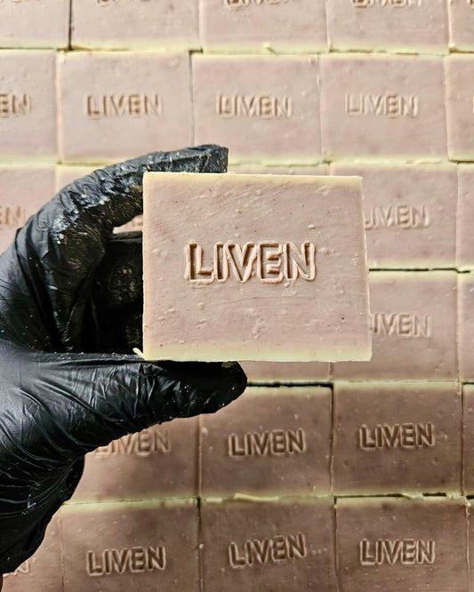 A hand wearing a black glove holds a rectangular bar of soap embossed with the brand name 'LIVEN' in the center. More identical soap bars are stacked in the background.