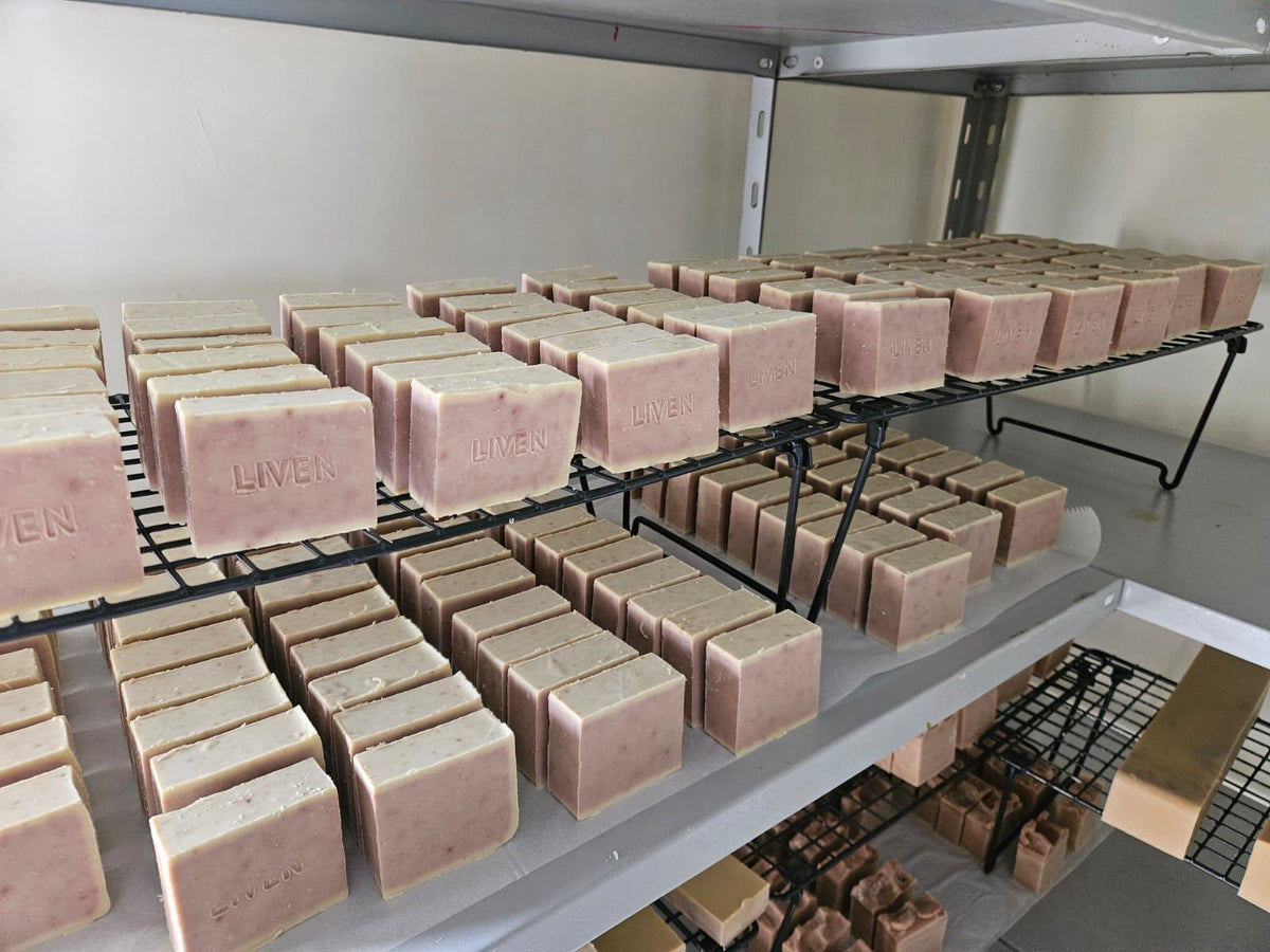 Multiple rectangular bars of soap in a dusty pink color arranged on black metal wire racks inside a storage room or curing space. The soap bars show the brand name 'LIVEN' embossed on the side.