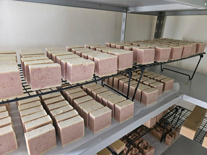 Multiple rectangular bars of soap in a dusty pink color arranged on black metal wire racks inside a storage room or curing space. The soap bars show the brand name 'LIVEN' embossed on the side.