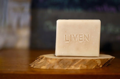 A rectangular bar of natural soap with grapeseed and olive oil, embossed with the word 'LIVEN' sits on a rustic wood slice coaster on a wooden table, with a blurred background.