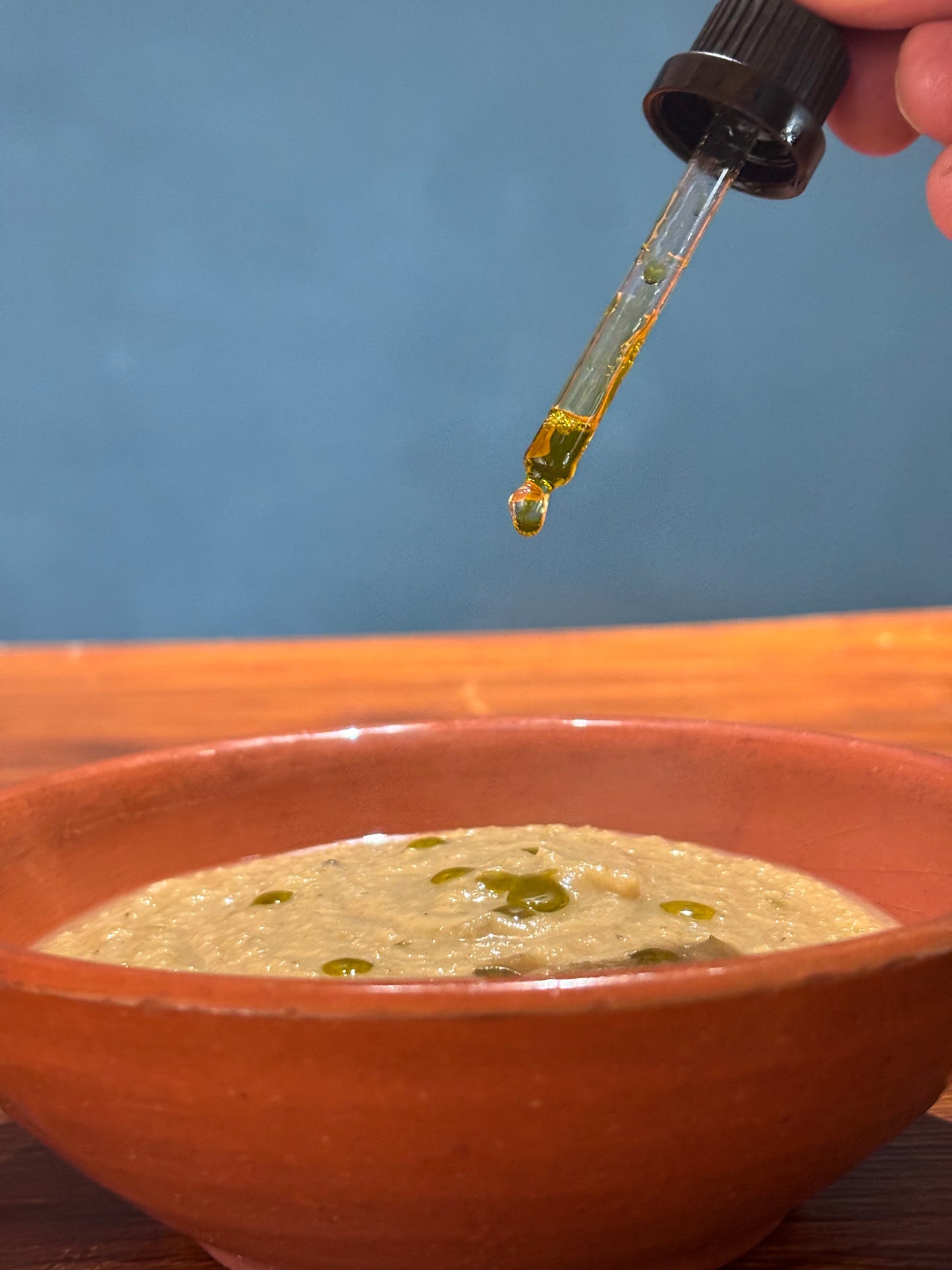 Dropper with oregano oil being added to a bowl of soup against a blue background