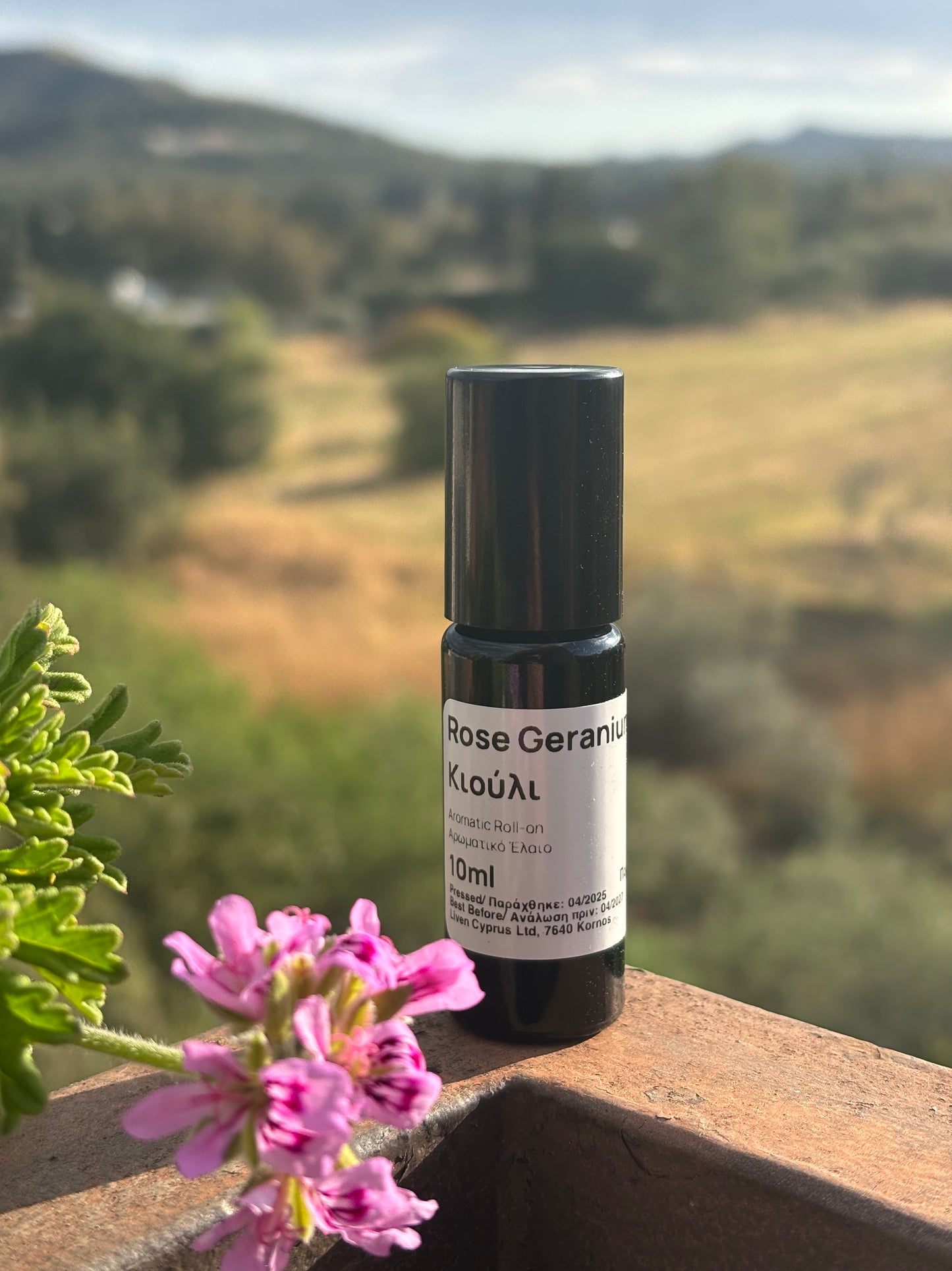 Rose geranium roll-on bottle with a mountain background, next to pink flowers