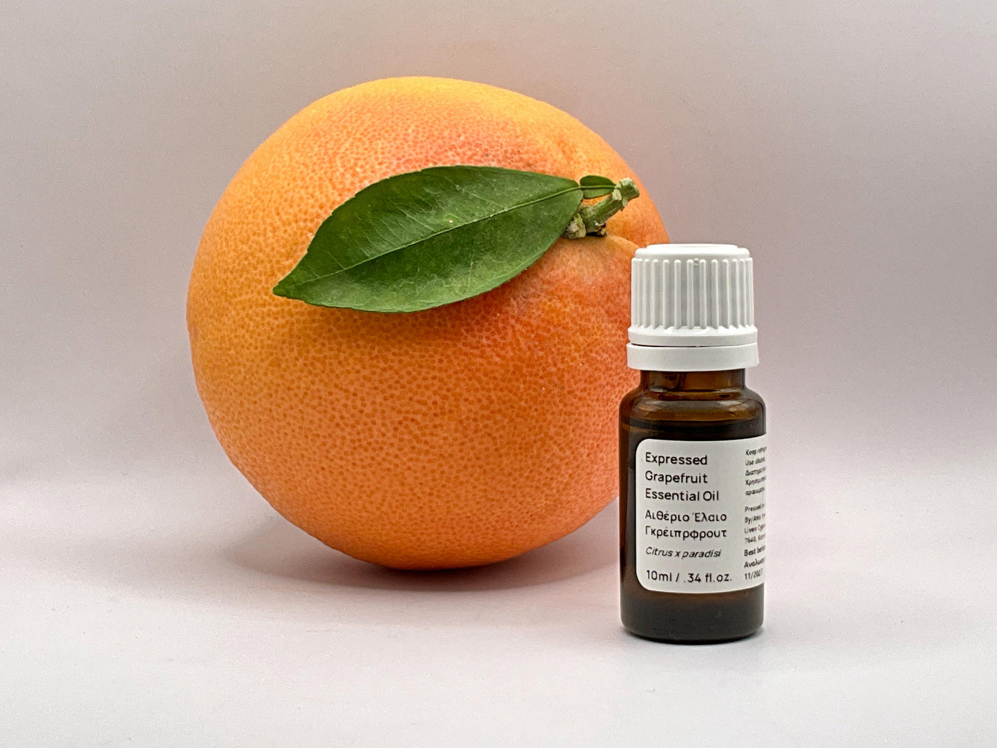 Grapefruit with a leaf next to a bottle of essential oil on a neutral background