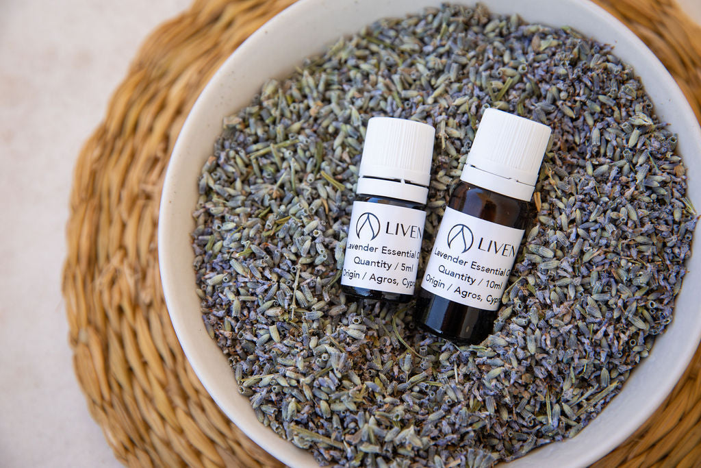 Two small bottles of essential oil placed on a pile of dried lavender flowers, within a bowl that is itself placed on a woven mat.